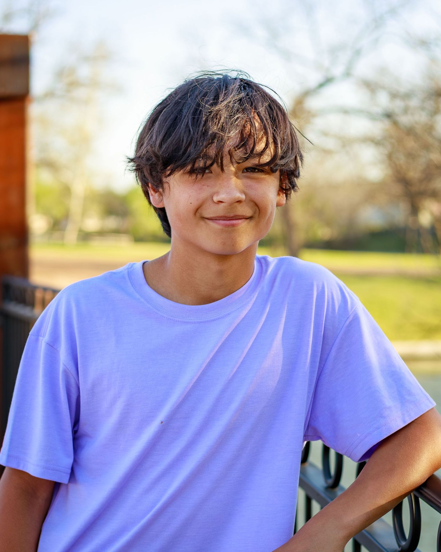 Smiling young boy with dark hair wearing a lavender t-shirt outdoors in a sunny park setting.