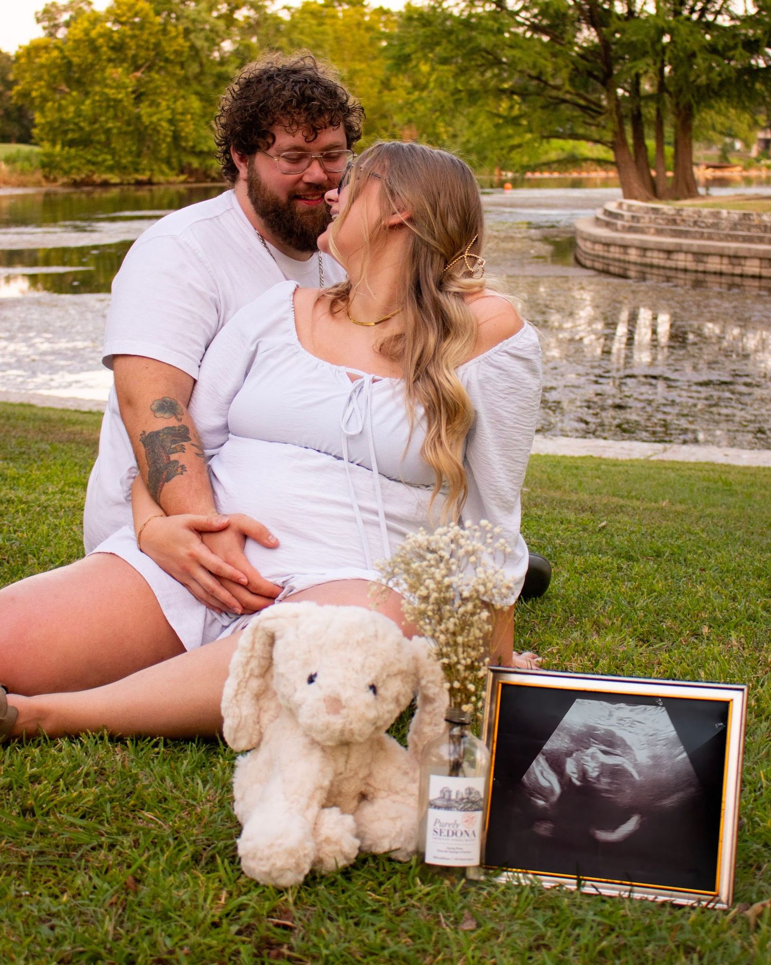 Couple sits on grass with stuffed animal and ultrasound photo during pregnancy announcement shoot.