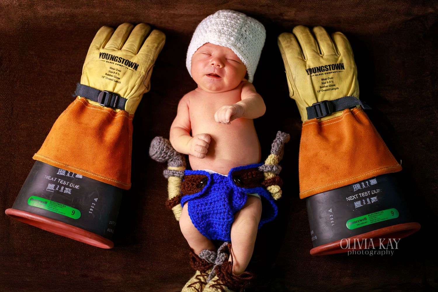 Newborn baby posed between large yellow firefighter gloves wearing a white hat and blue crocheted diaper cover.