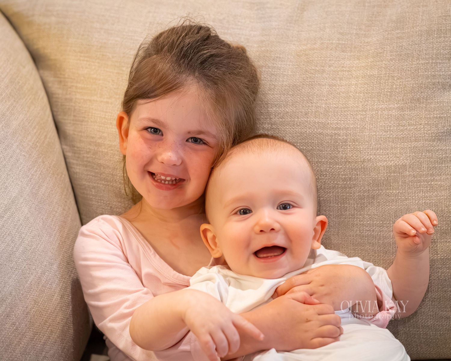 Smiling toddler girl in pink hugging a happy laughing baby on a gray couch.