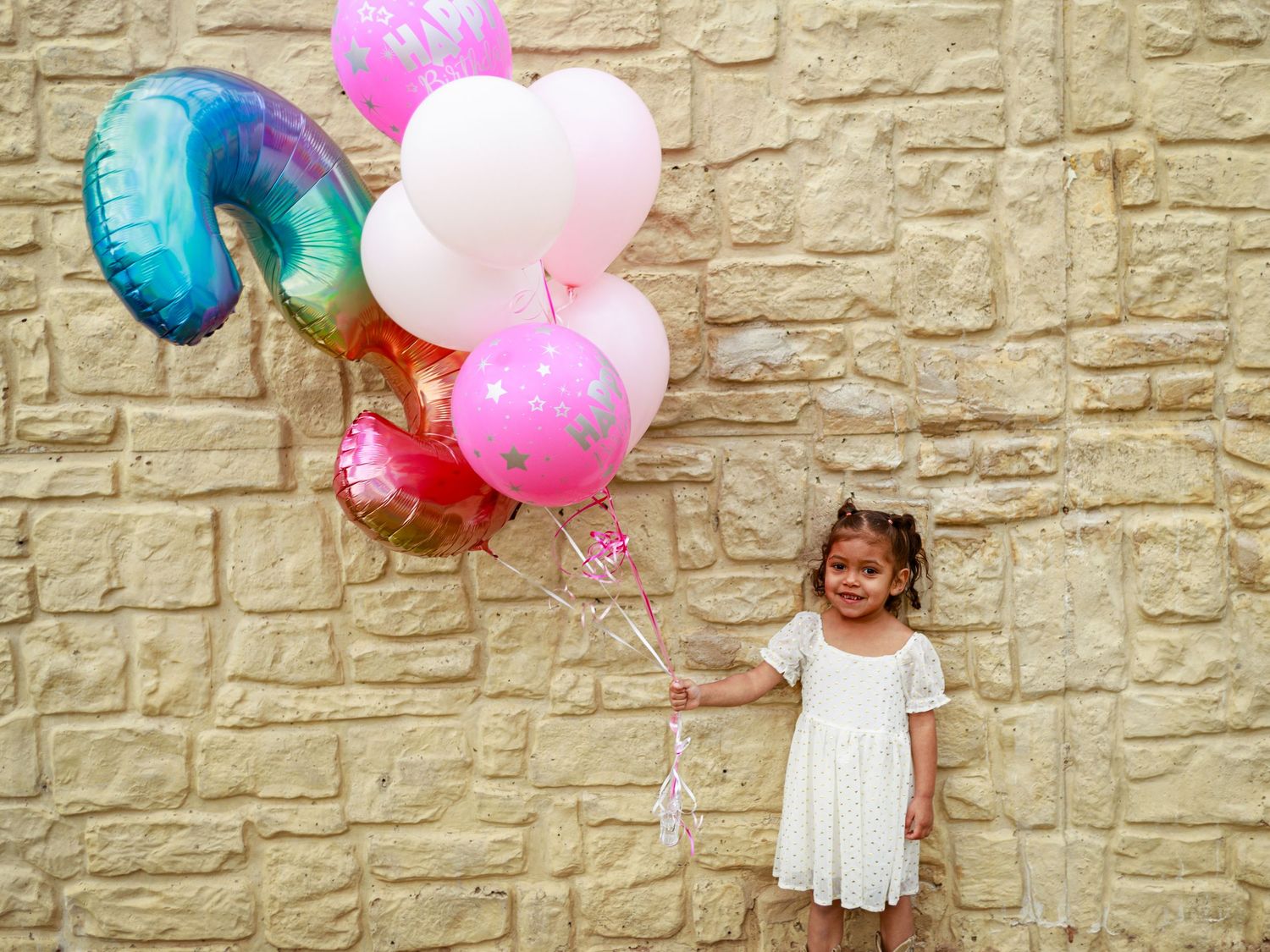 Young girl in white dress holding colorful birthday balloons against a stone wall backdrop.