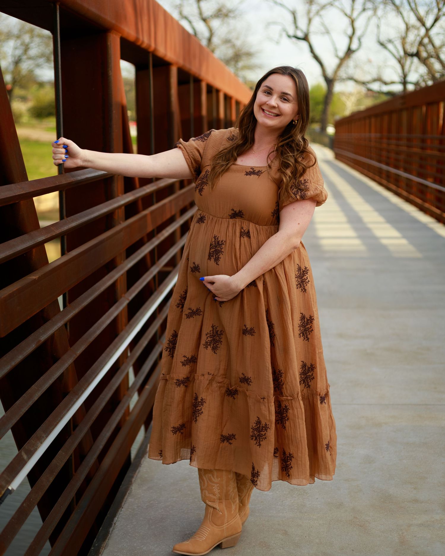 Someone in a brown floral dress and boots poses on a wooden bridge walkway during golden hour.