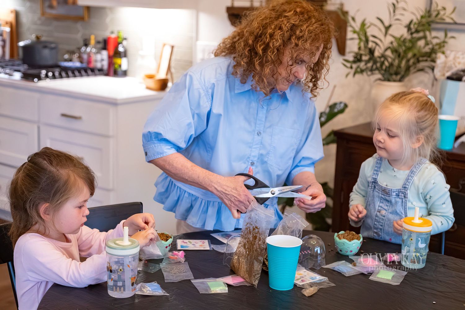 Woman and two young girls doing crafts at kitchen table with scissors, jars, and colorful supplies.