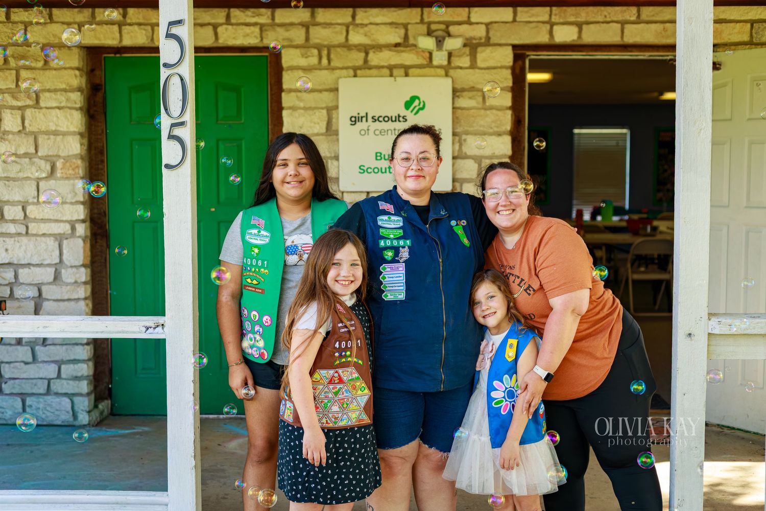 Five Girl Scouts and leaders pose smiling outside a stone building with green doors marked 505.