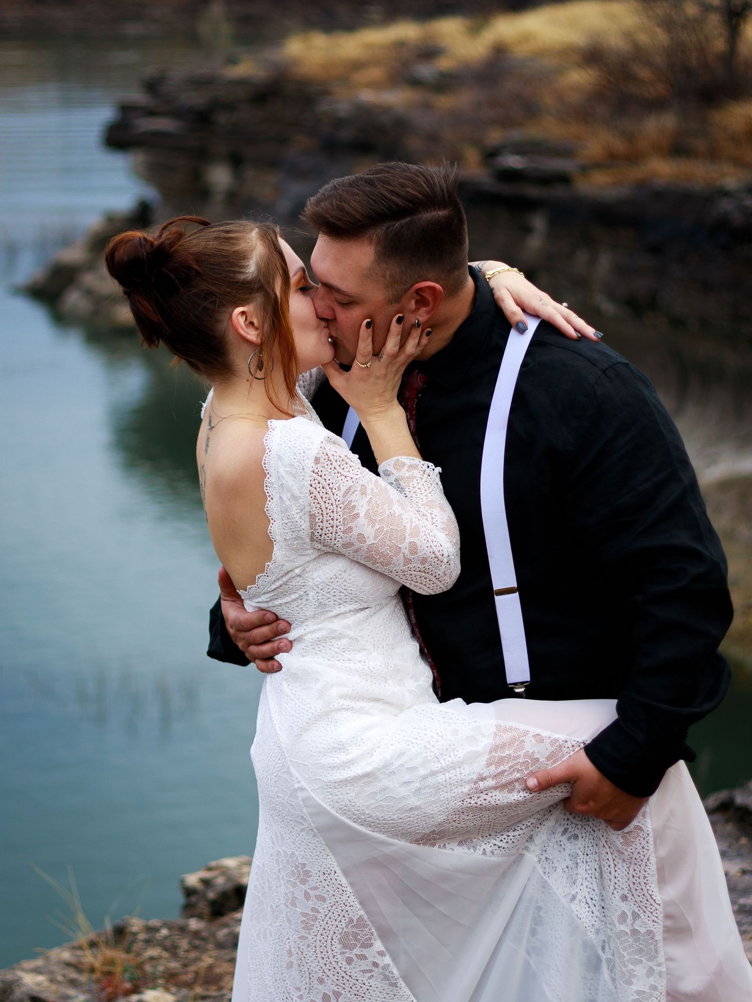 Bride and groom sharing a romantic kiss outdoors near a lake, bride in lace dress, groom in black suit with white suspenders.