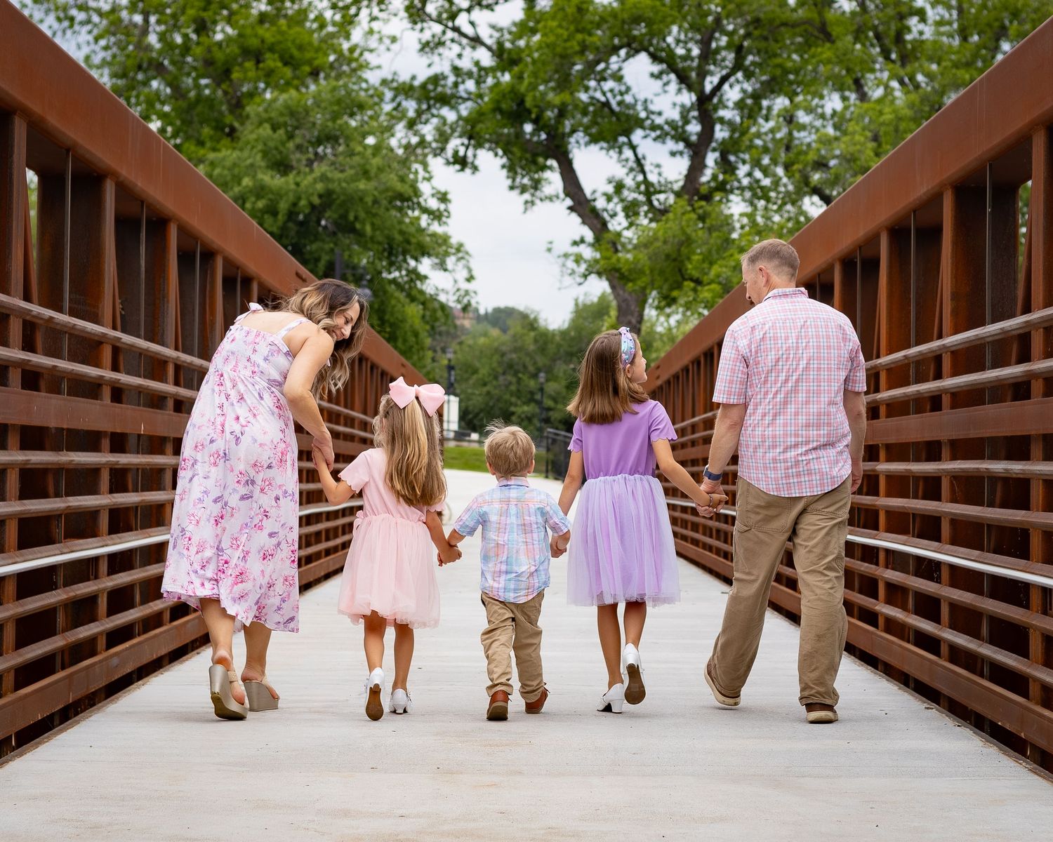 Family of five walking hand-in-hand across a scenic bridge, dressed in coordinating pastel outfits, surrounded by lush greenery.