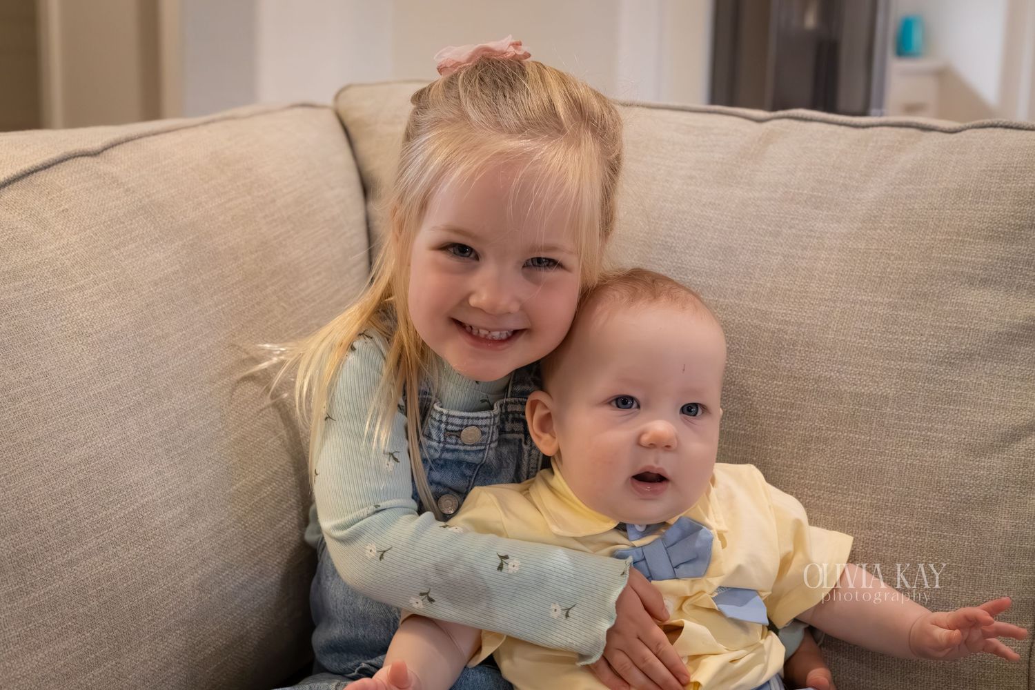 A smiling toddler girl with blonde hair holds a baby boy on a gray couch.