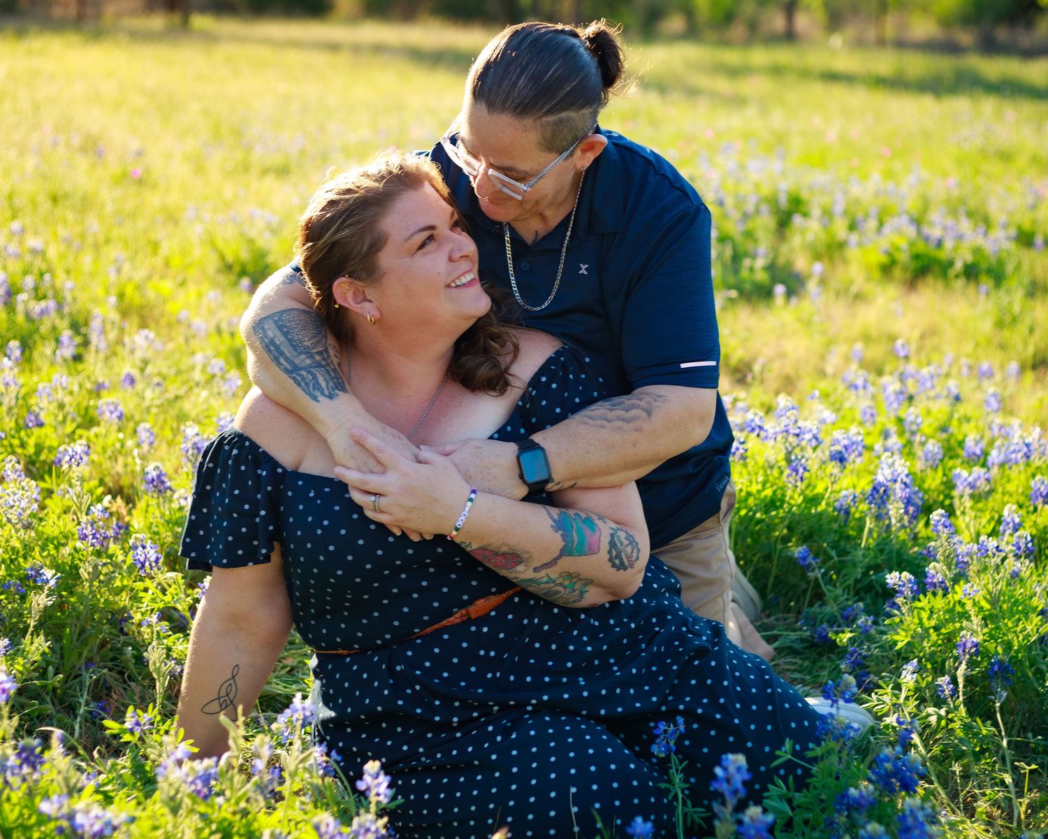 Couple embracing in a sunny bluebonnet field, woman sitting in polka dot dress, man hugging her from behind.