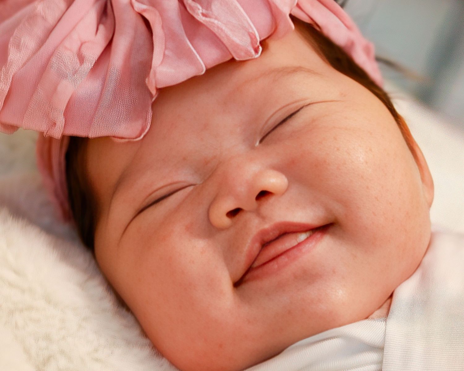 Smiling sleeping baby girl wearing a pink flower headband, peacefully resting on a white blanket.