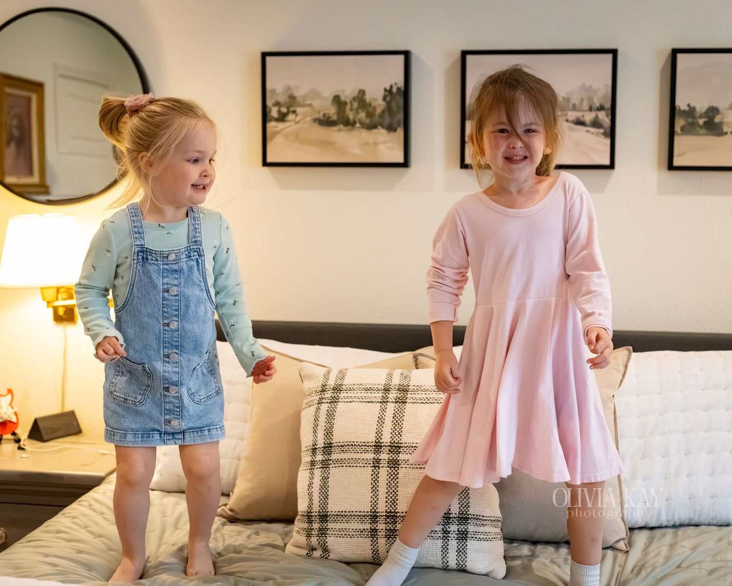 Two young girls in dresses joyfully jumping on a bed in a cozy decorated bedroom.