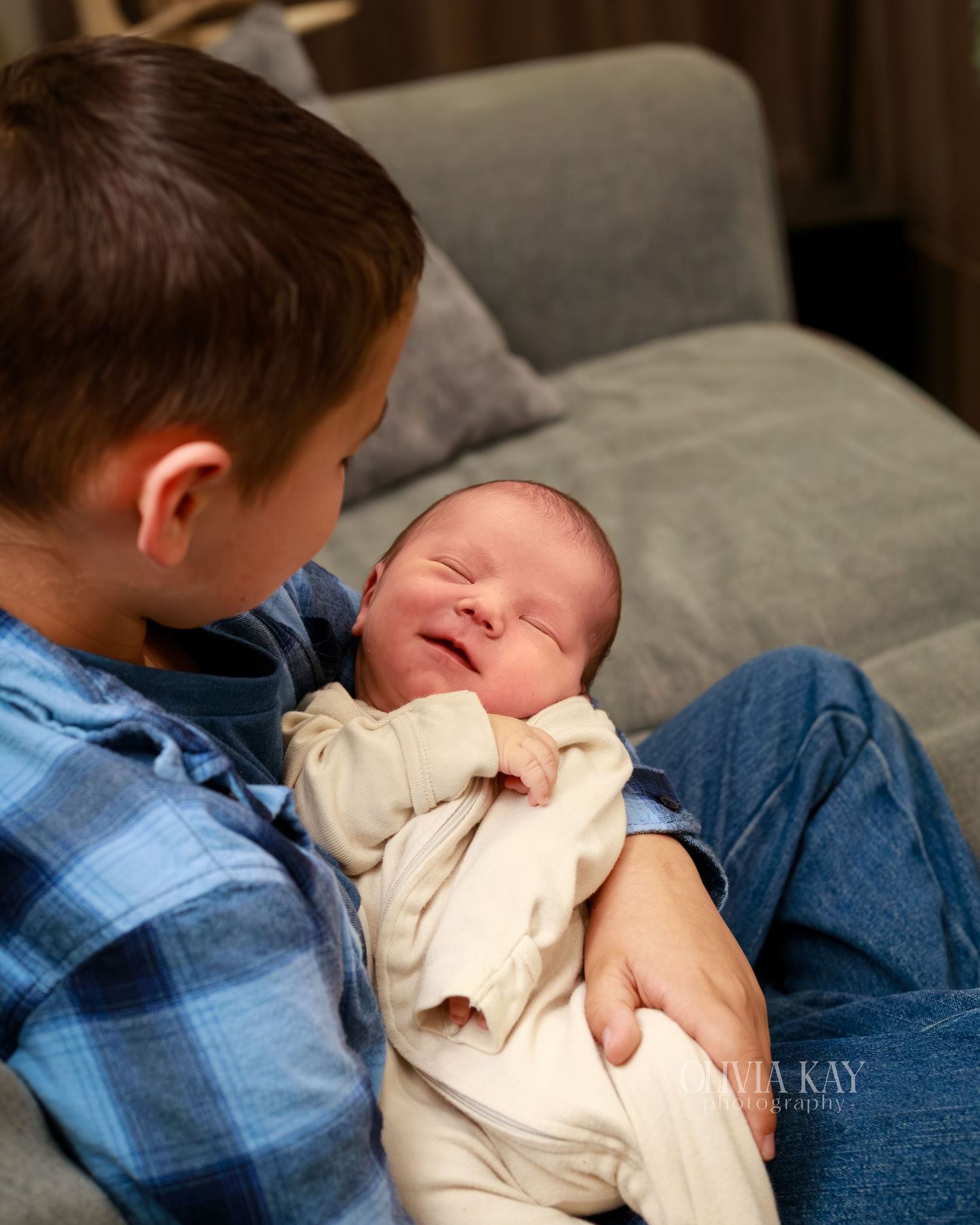 Older boy in blue plaid shirt holding smiling newborn baby sibling on gray couch.