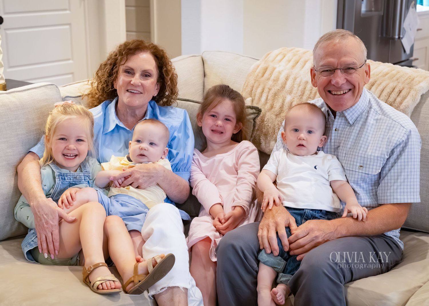 Grandparents sitting on a couch smiling with four young grandchildren, including two toddlers and two babies.