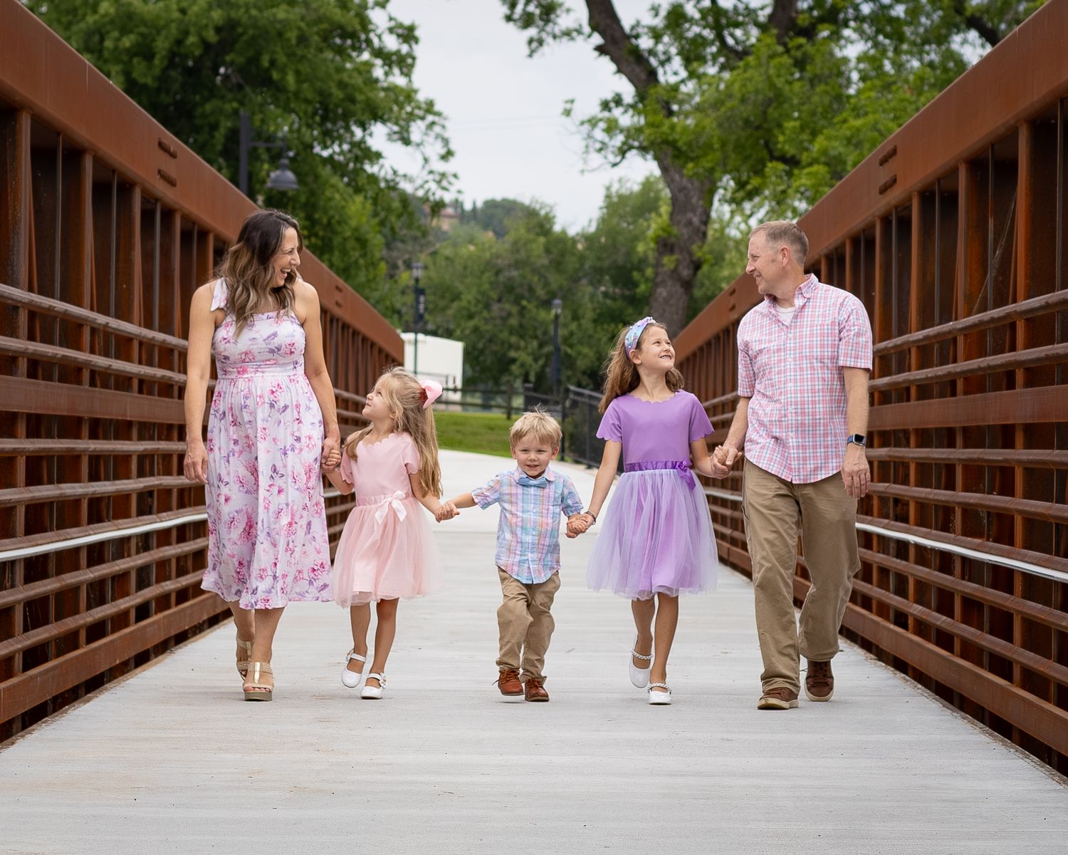 Family of five walking hand-in-hand on a wooden bridge, wearing pastel outfits, surrounded by lush green trees.
