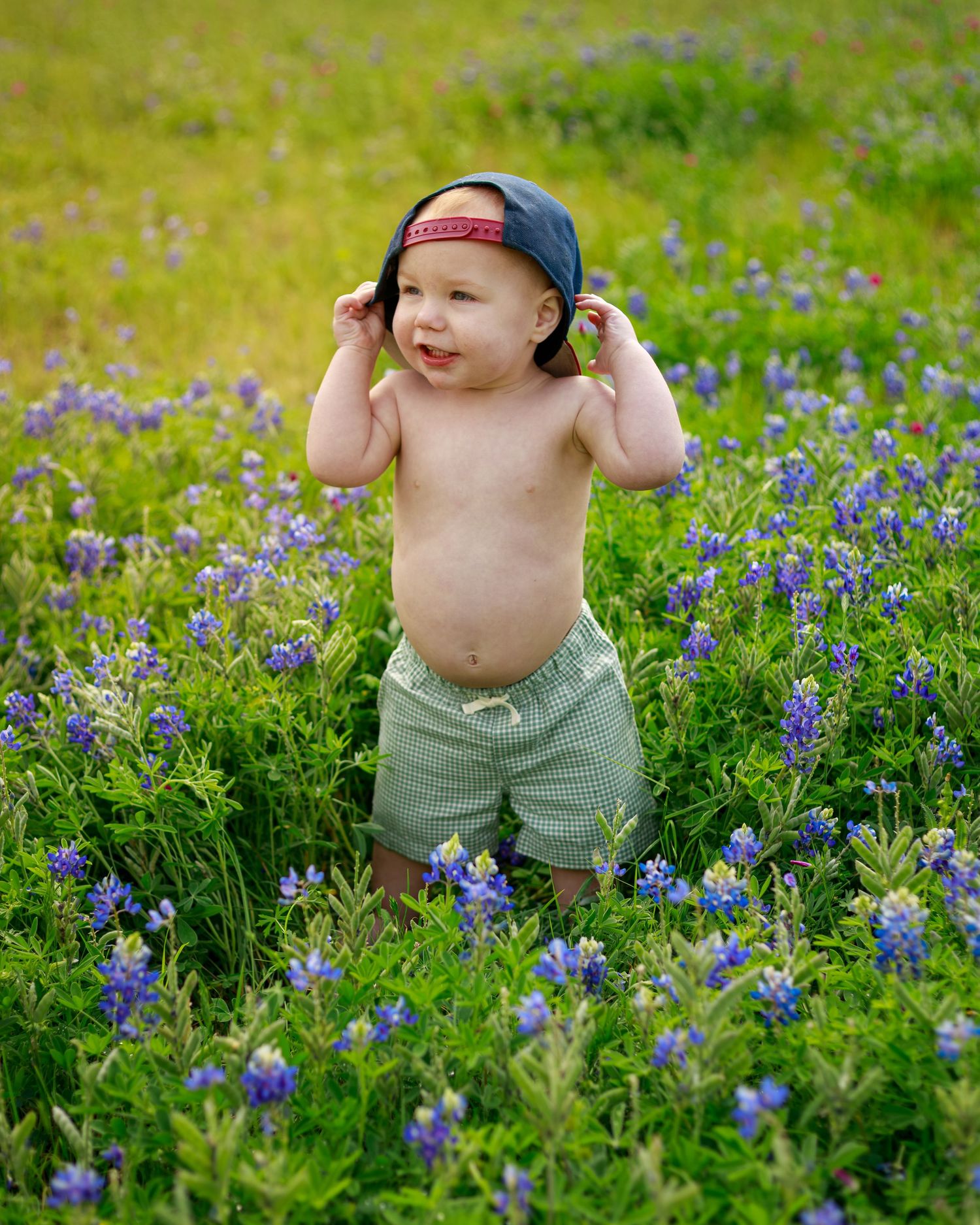 A shirtless baby in green shorts and a backwards cap stands smiling in a field of purple bluebonnet wildflowers.