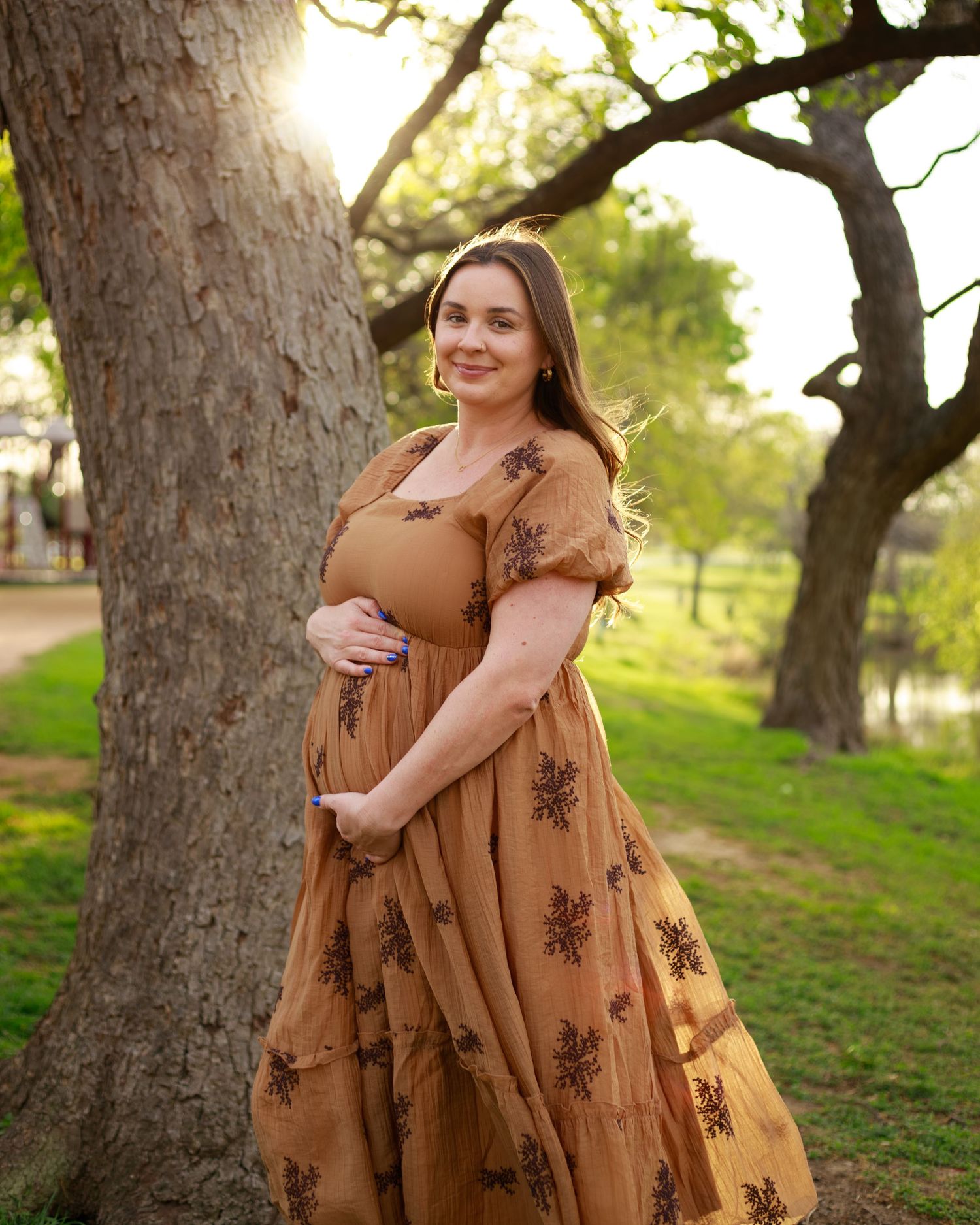 A person wearing a flowing brown dress with floral embroidery poses against a tree in a sunlit outdoor setting.