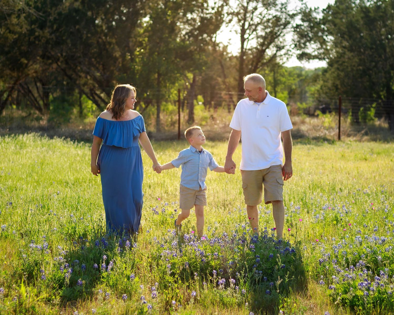 Family of three holding hands in a sunny wildflower meadow, mother in blue dress, father in white shirt, young boy between them.