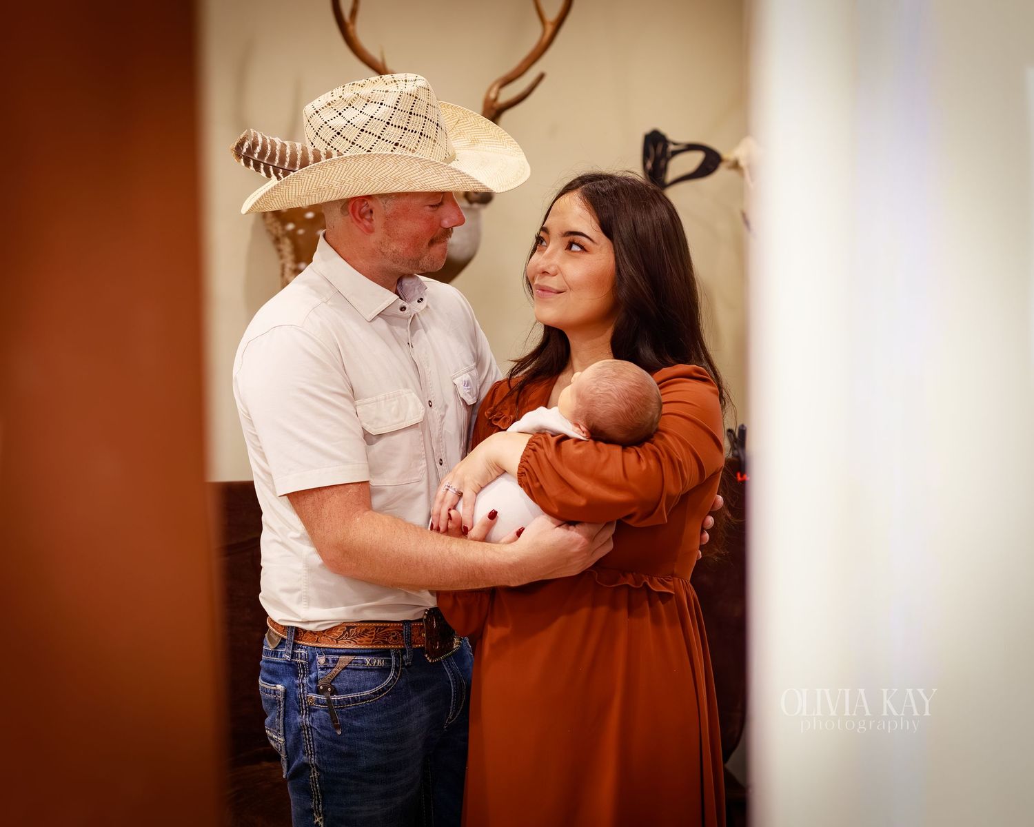 A couple in western attire holding their newborn baby, smiling warmly in a rustic indoor setting.