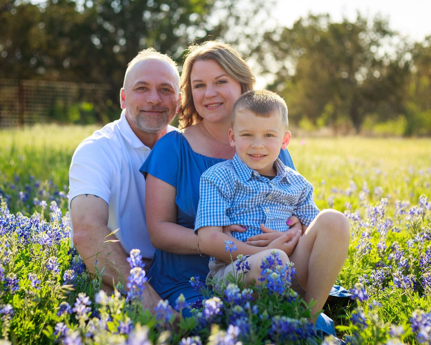 Family of three smiling in a field of bluebonnets on a sunny day in Texas.