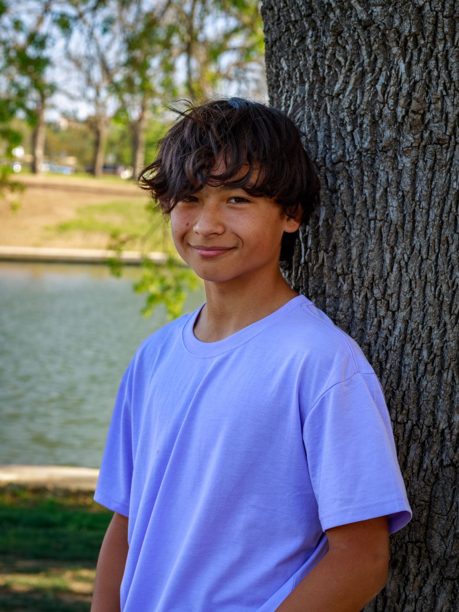 Smiling teenage boy with curly dark hair wearing a lavender t-shirt, leaning against a tree near a lake.