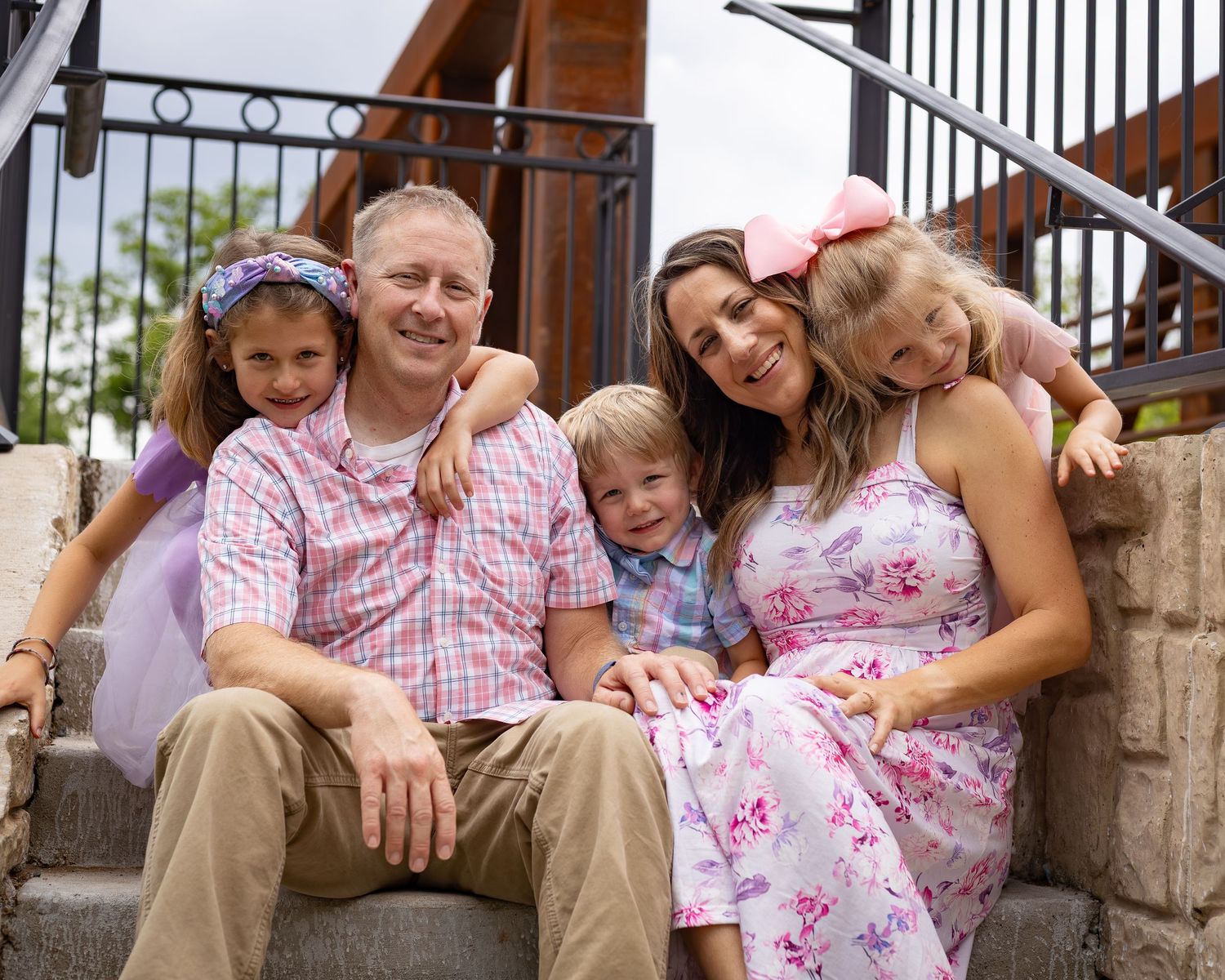 Happy family of five smiling together on outdoor steps, wearing coordinating pink and floral outfits on a sunny day.