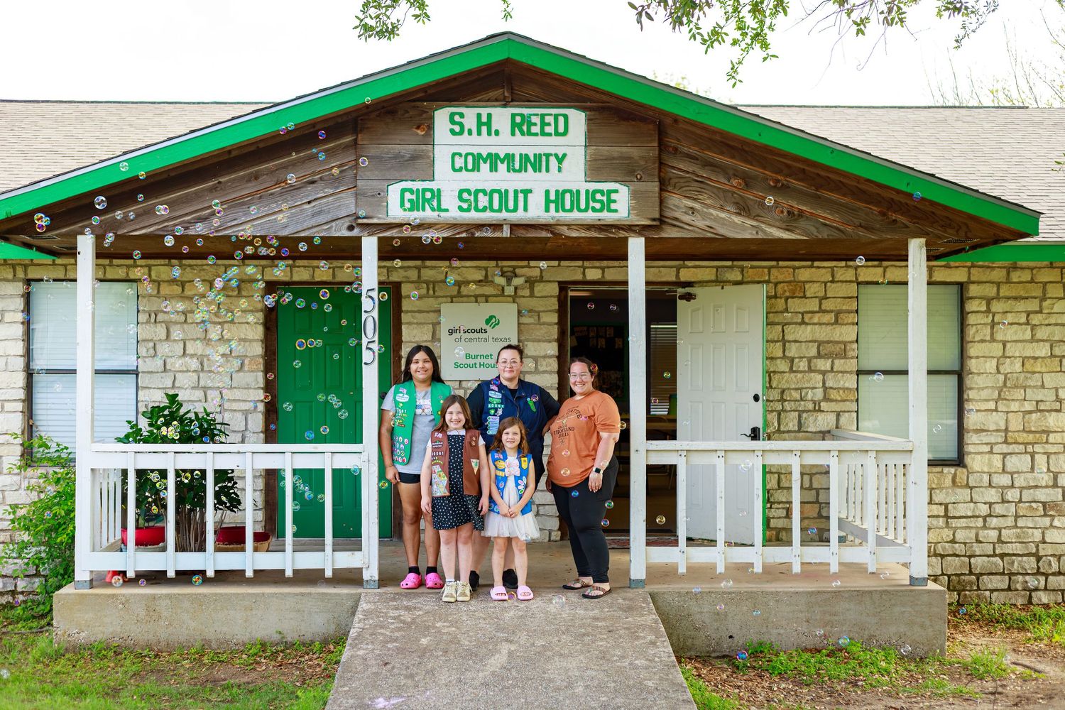 Family posing in front of S.H. Reed Community Girl Scout House, a stone building with green trim and white sign.