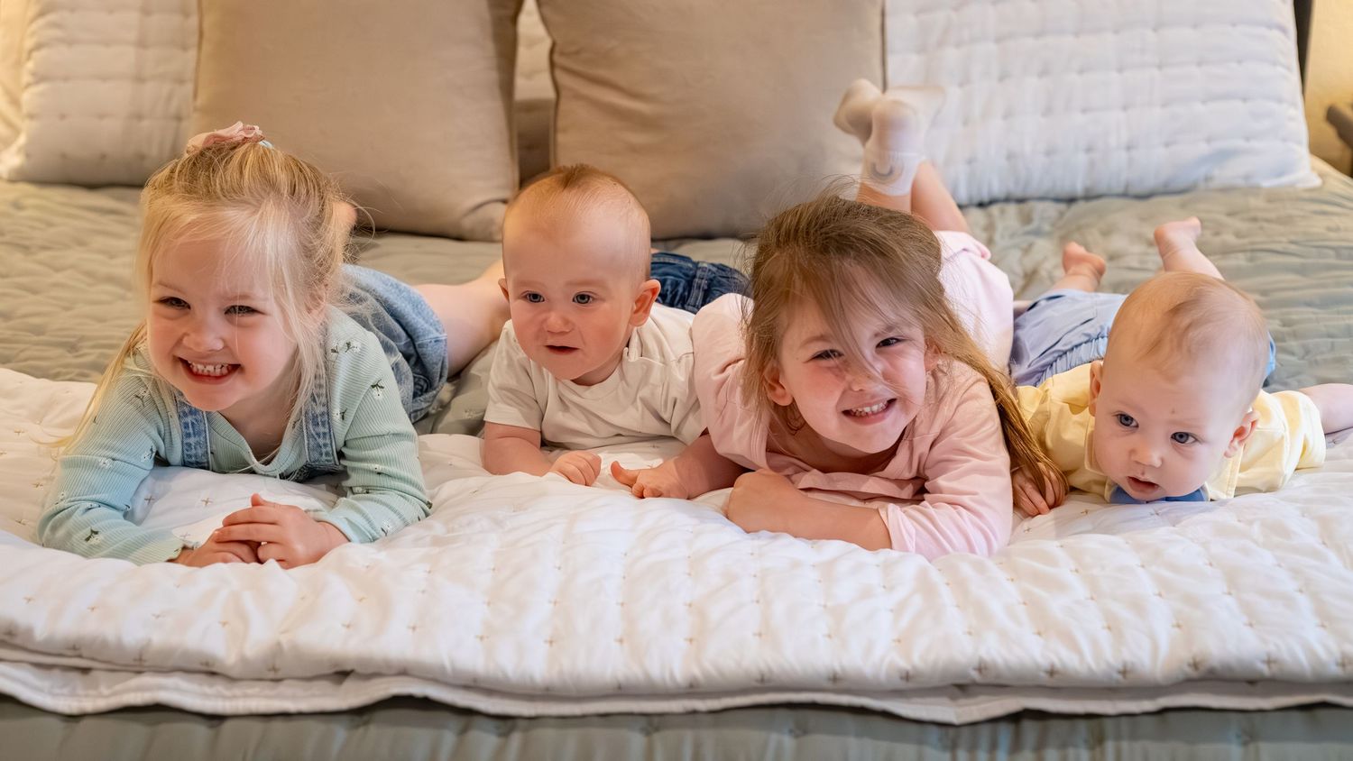 Four adorable babies lying on a couch together, smiling and posing for a cute group photo.