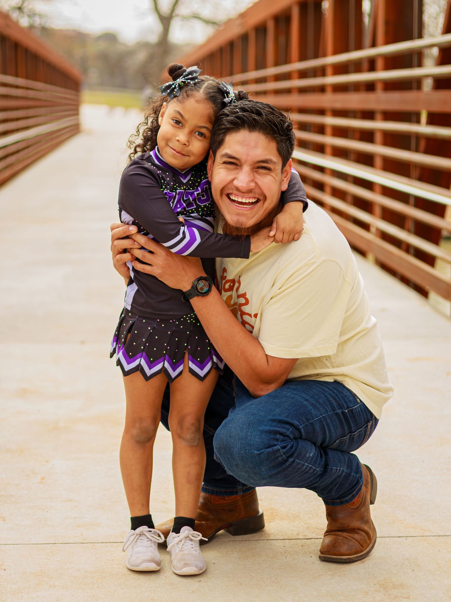 A smiling man crouches on a bridge hugging a young girl in a black and purple cheerleading uniform.