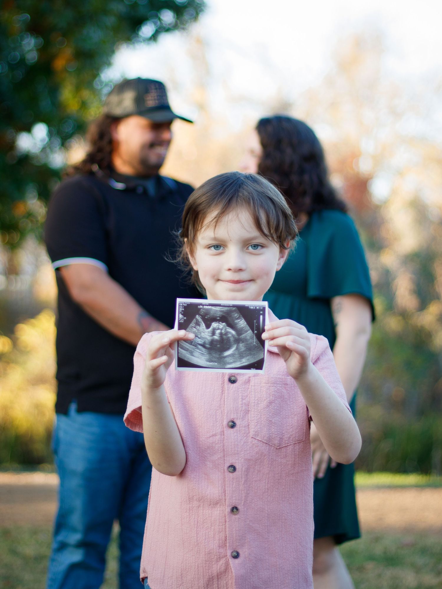 A child in a pink shirt holds up an ultrasound photo while standing outdoors.