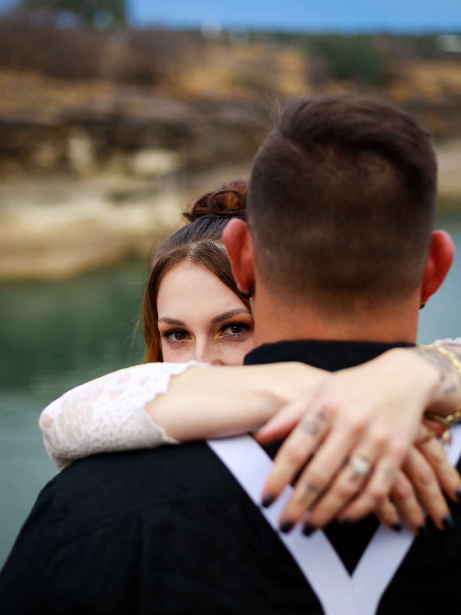 Bride in lace wedding dress embracing groom from behind near a lake, showing diamond engagement ring on her hand.