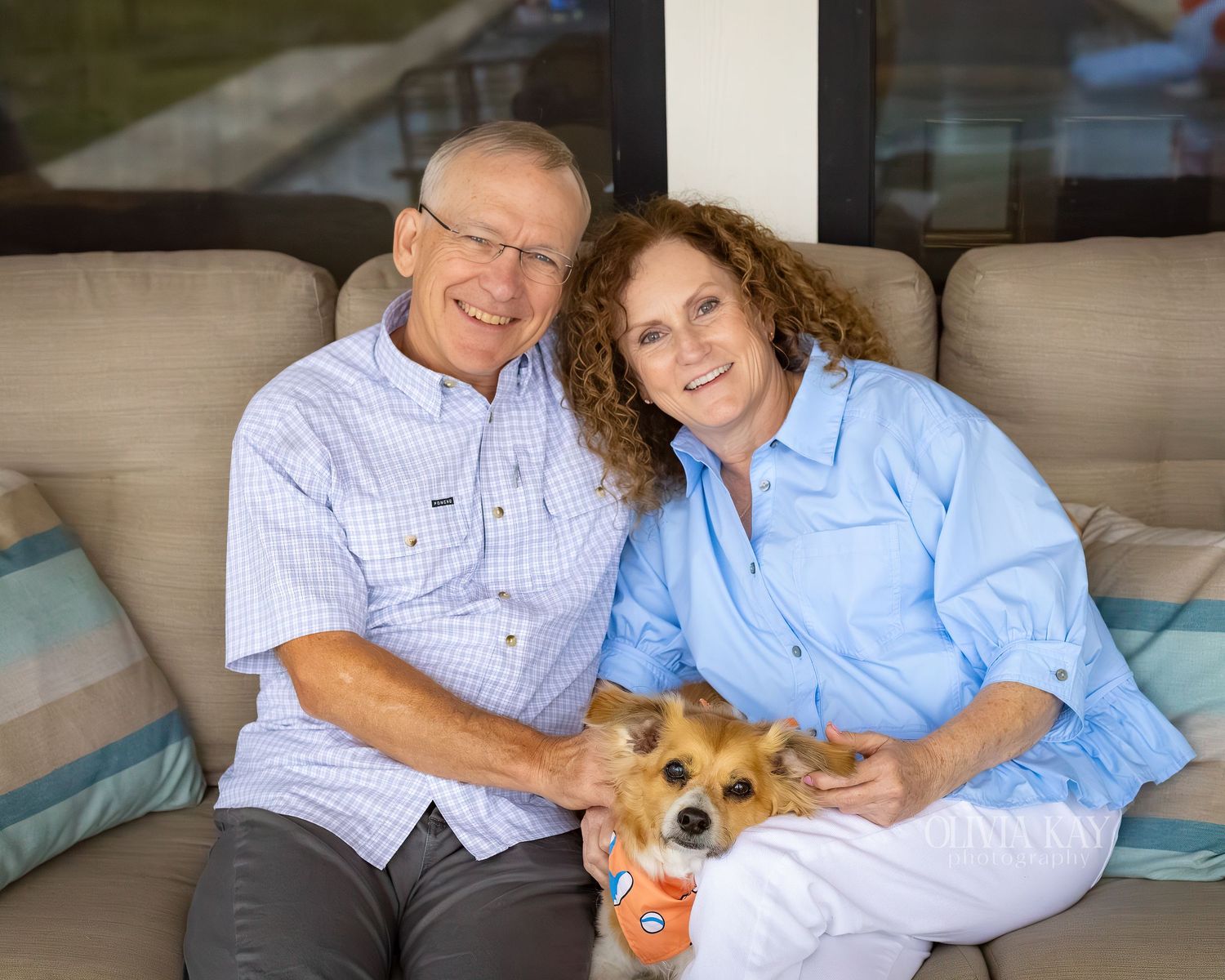 Smiling couple sitting on a couch with a small Corgi dog, dressed in light blue shirts in a cozy outdoor setting.