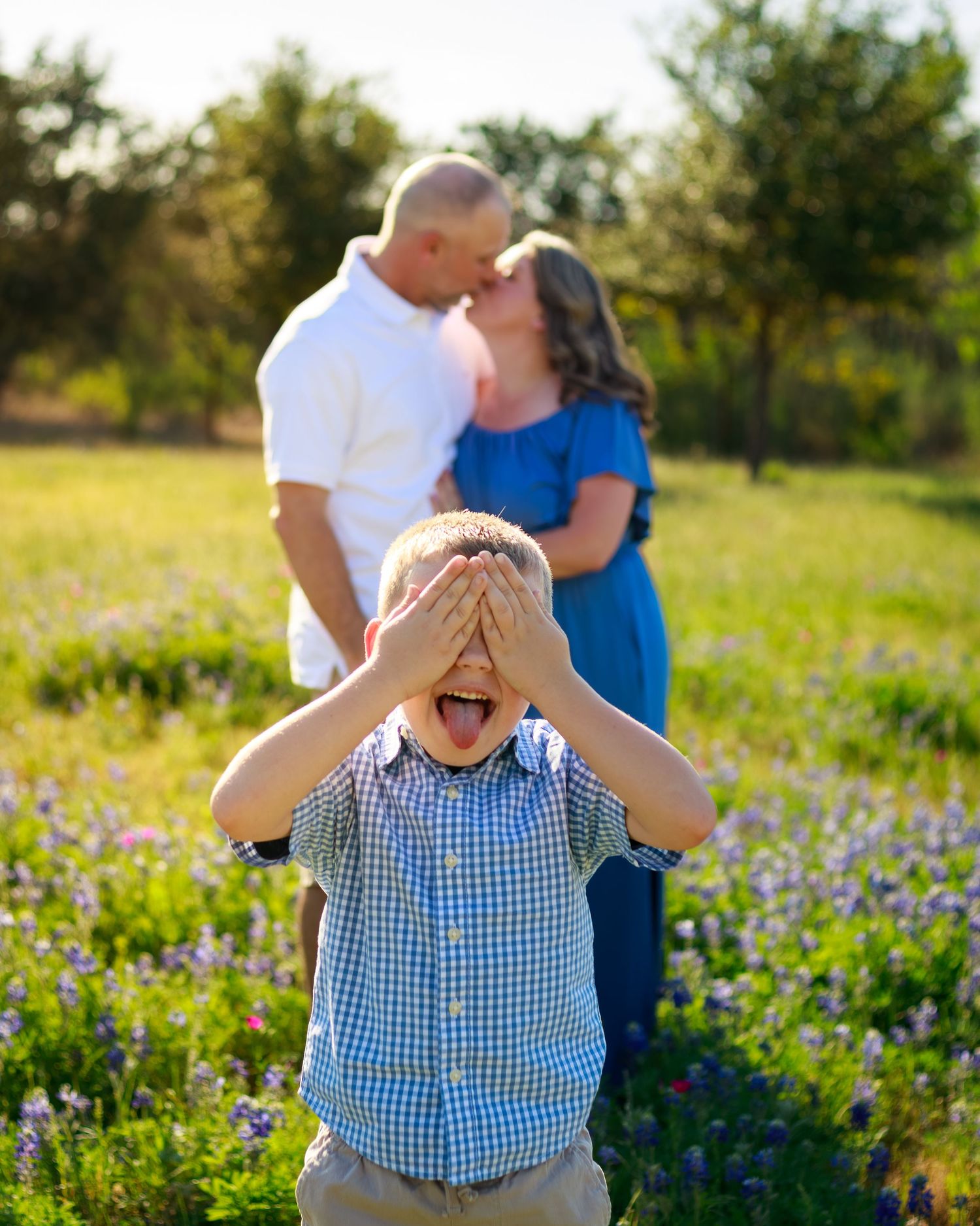 A young boy covers his eyes laughing as his parents share a romantic kiss in a sunny wildflower field behind him.