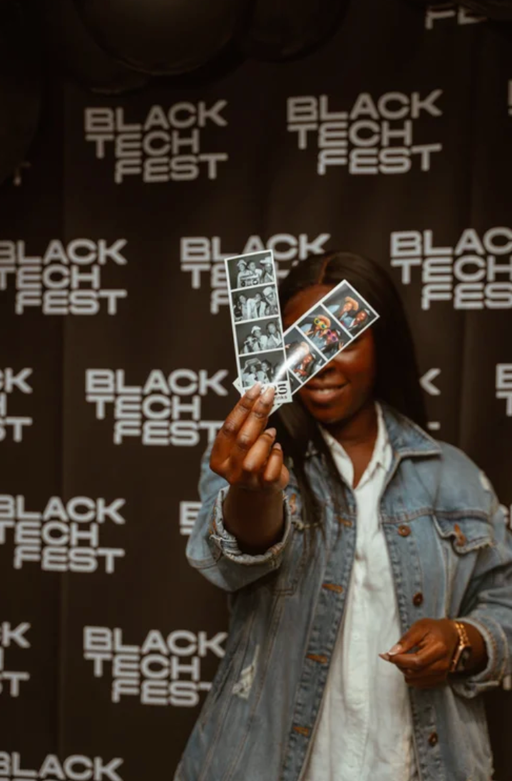 Person in denim jacket holds up cards against Black Tech Fest branded backdrop.
