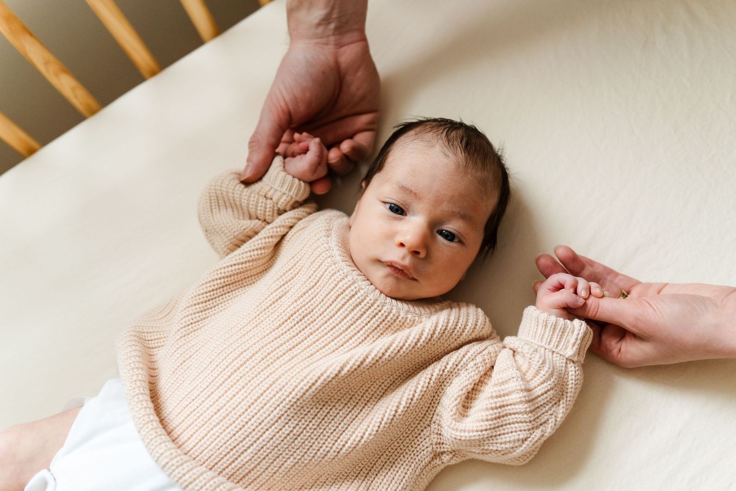A baby wearing a ribbed beige sweater lies on white bedding while hands reach to comfort.