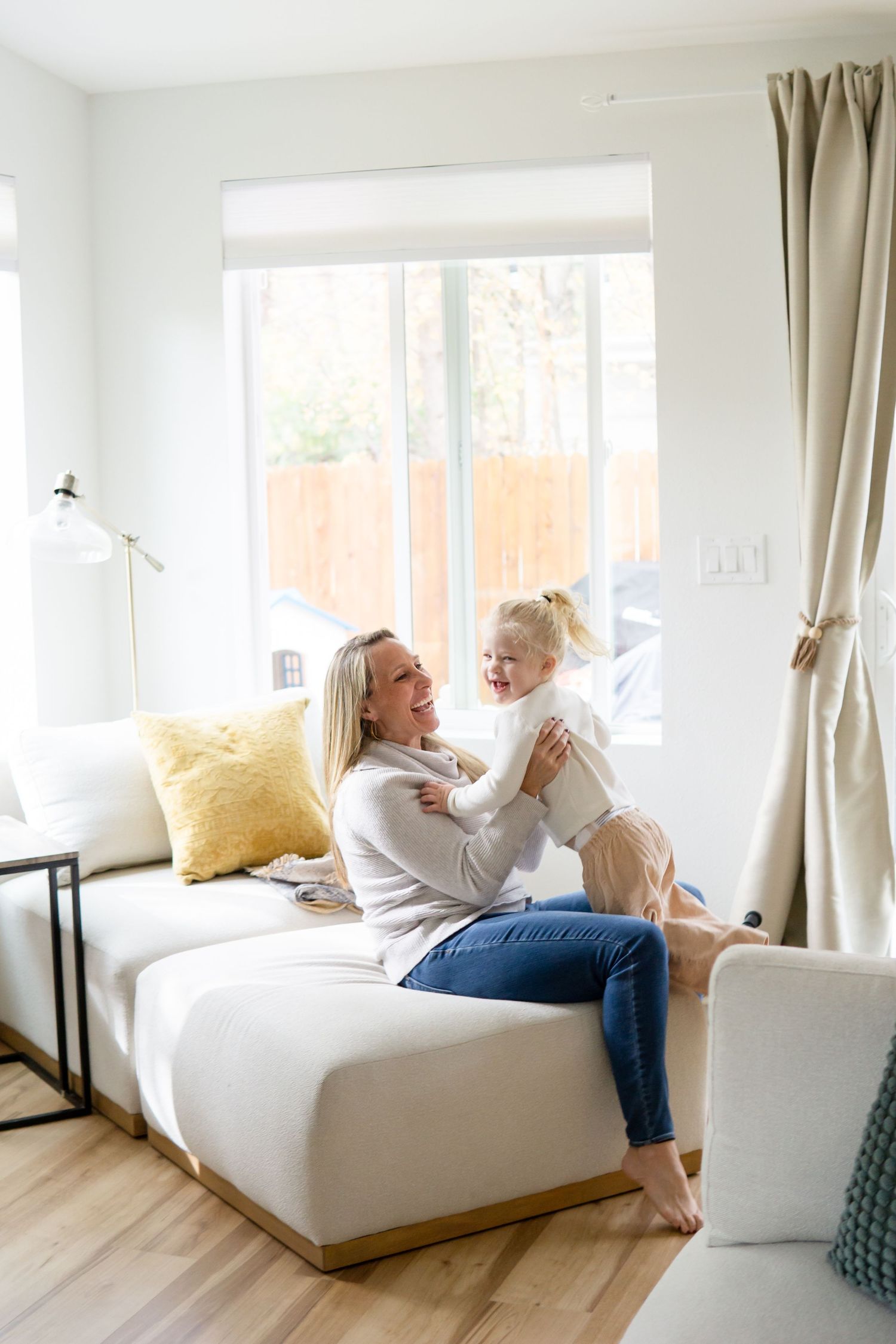 A bright living room interior with white couch, yellow pillow, and natural light streaming through windows.