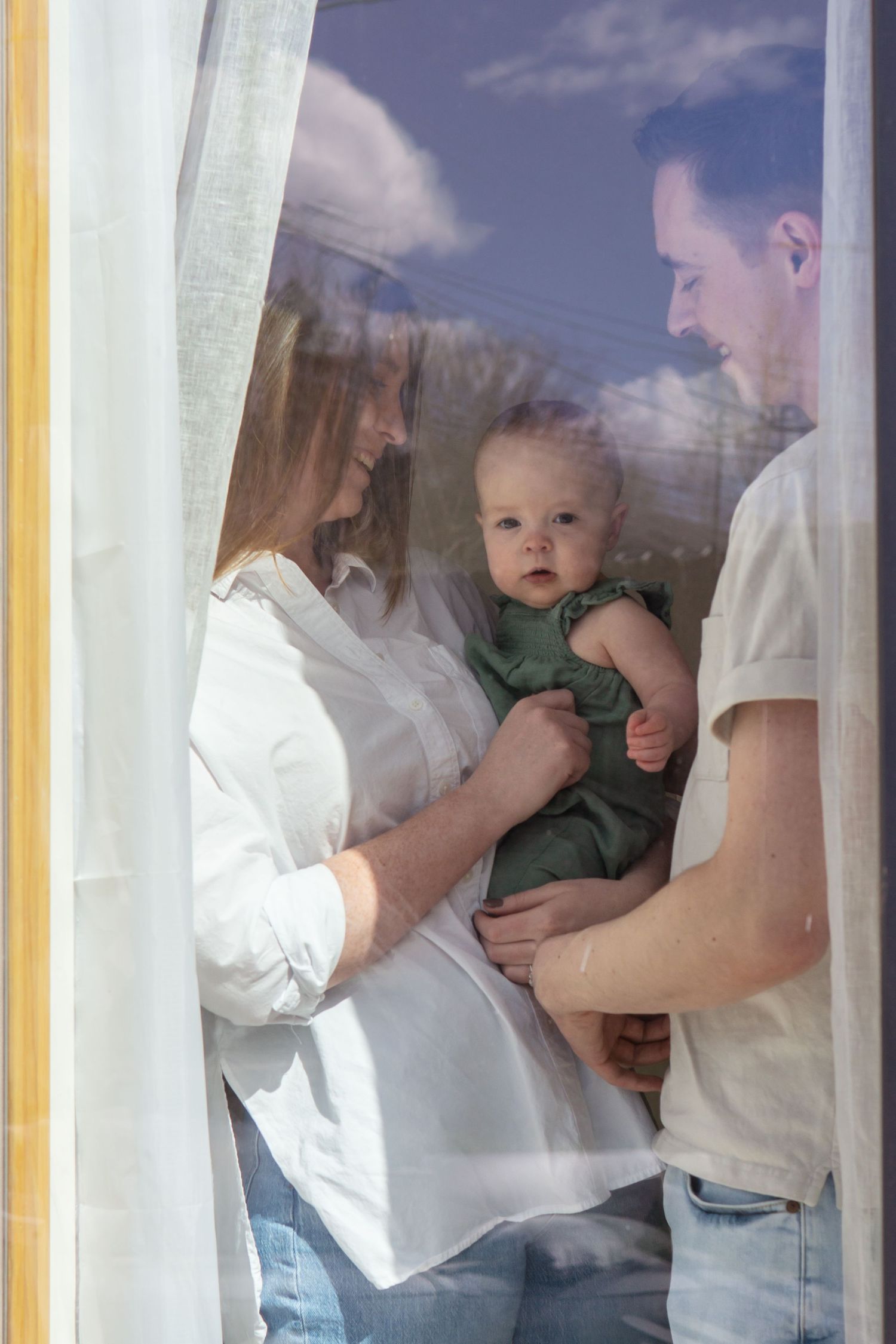 A baby in green clothing is held by parents behind sheer curtains near a window.