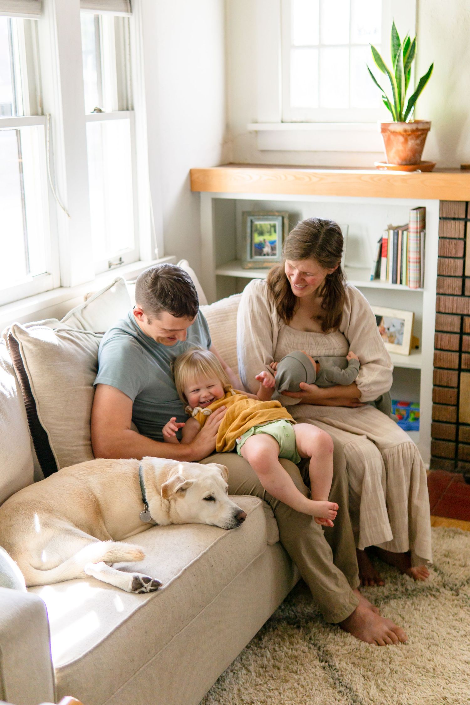 Family relaxing on beige couch with dog in sunlit room with plants.