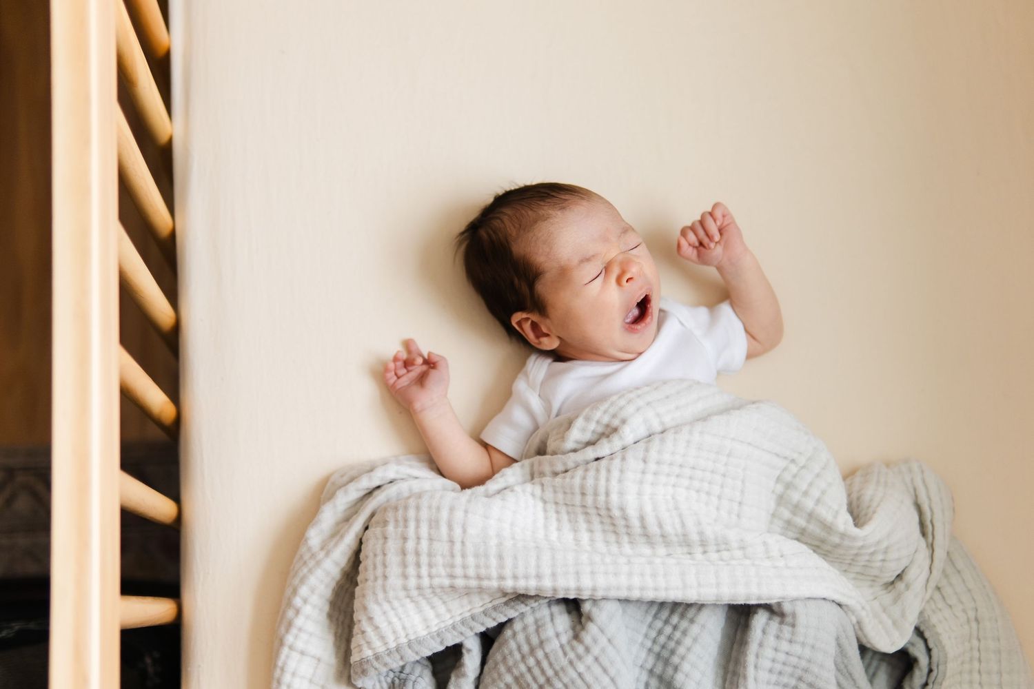 A sleeping newborn wrapped in a white blanket lying against a neutral wall.