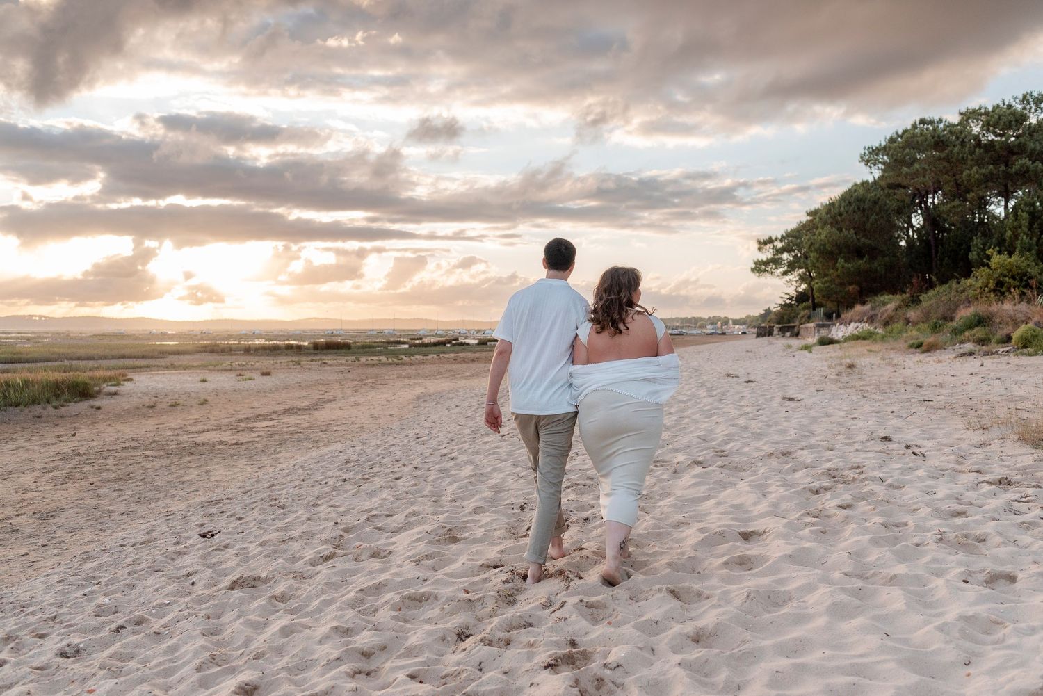 Couple walking hand in hand along a sandy beach during golden hour with boats in the distance.
