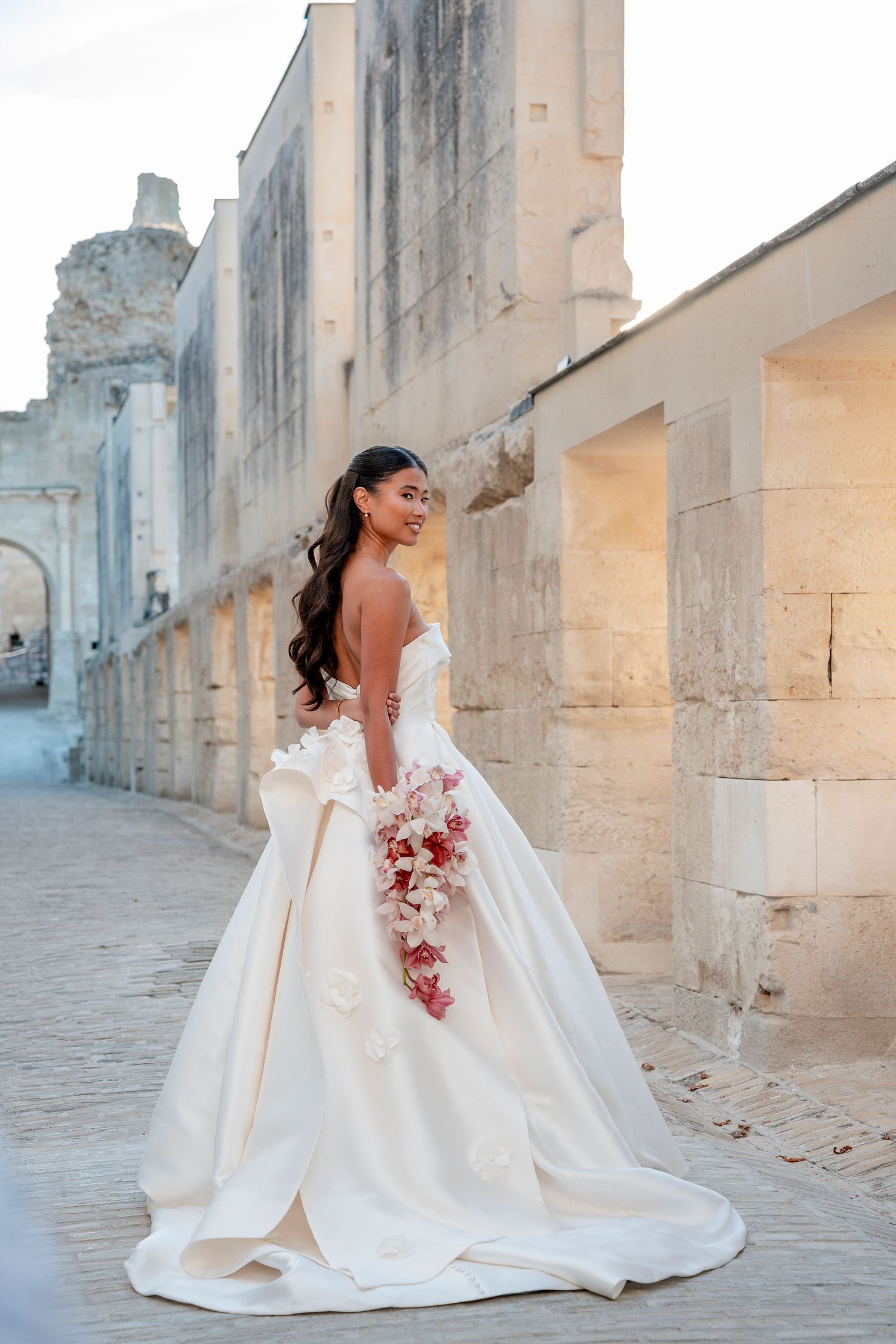 Bride in an elegant white ball gown holding a pink and red bouquet while posing on a stone path near ancient limestone walls.