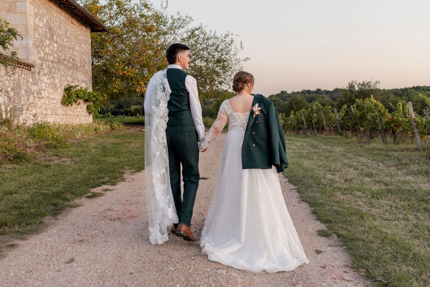 Bride in a white gown and groom in a green vest and trousers walk hand in hand down a path beside a vineyard at sunset.