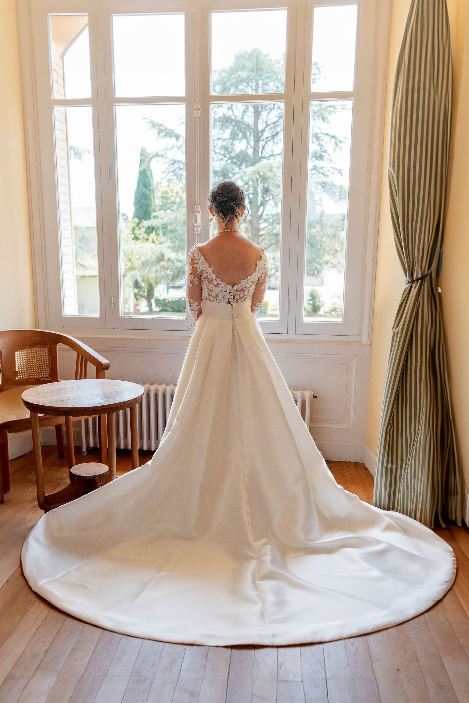 Elegant wedding dress with long train displayed in sunlit room with white curtains.