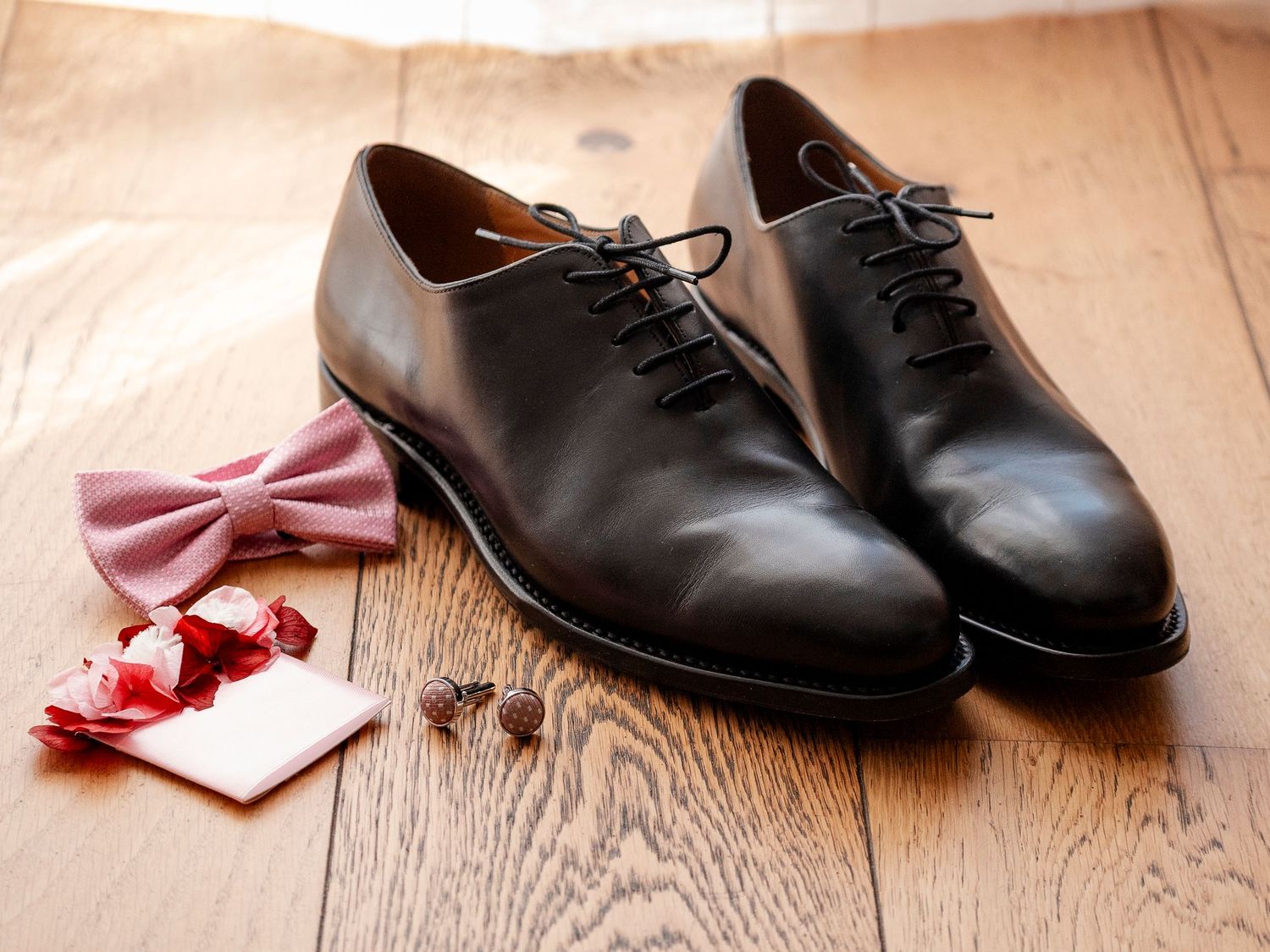 Black leather dress shoes and pink bow tie arranged on wooden floor for formal occasion.