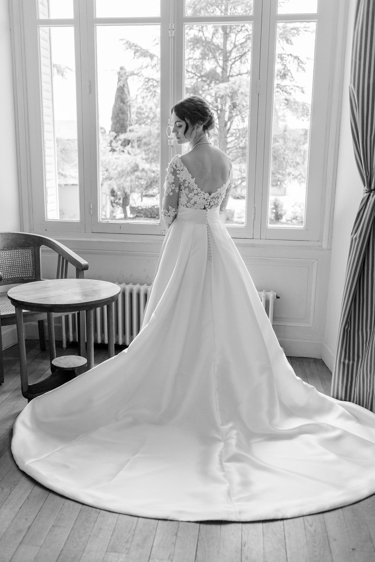 Elegant white wedding dress with long train displayed in sunlit room with wooden floors.