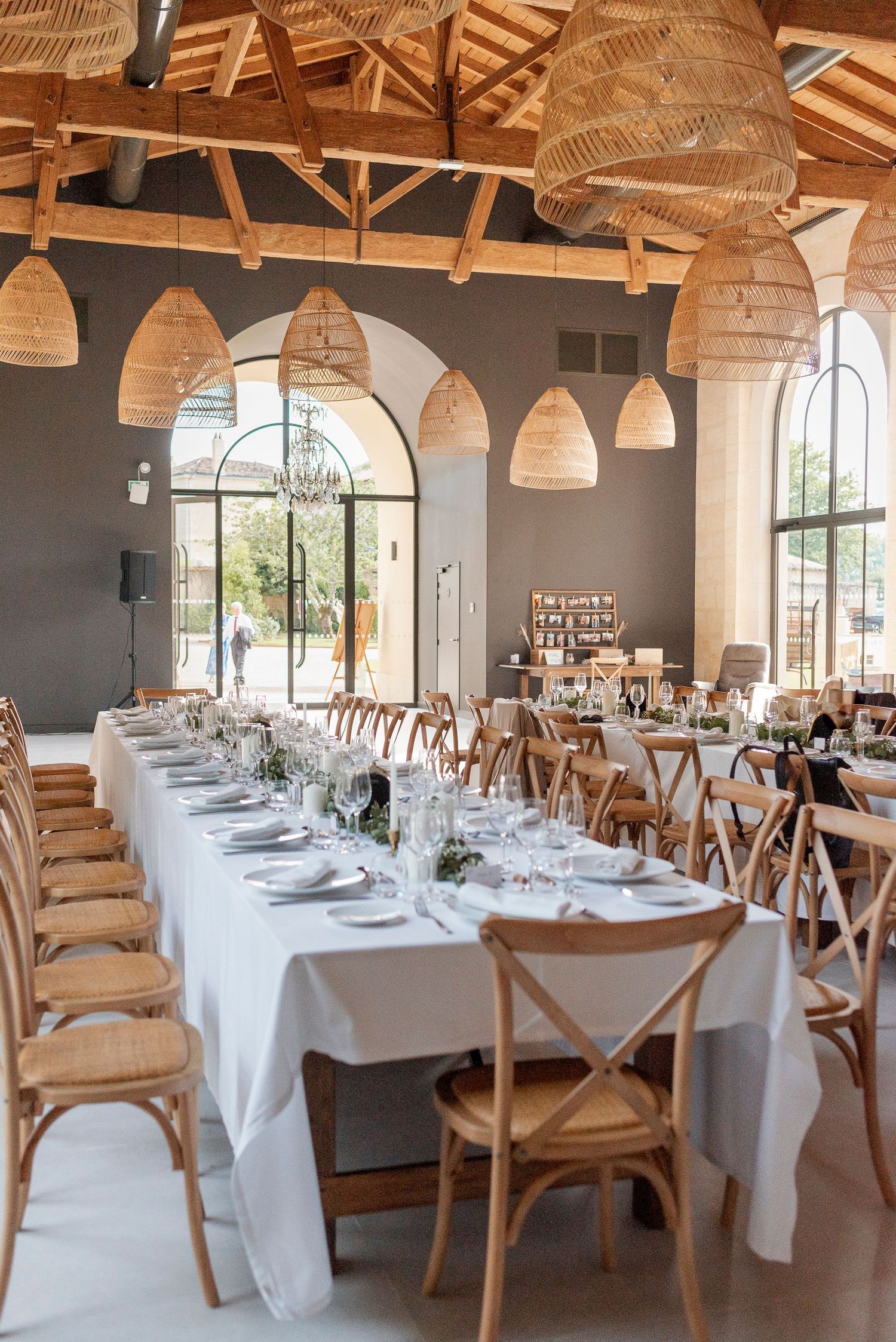 Reception venue with wooden crossback chairs and white tablecloths under rattan pendant lights.