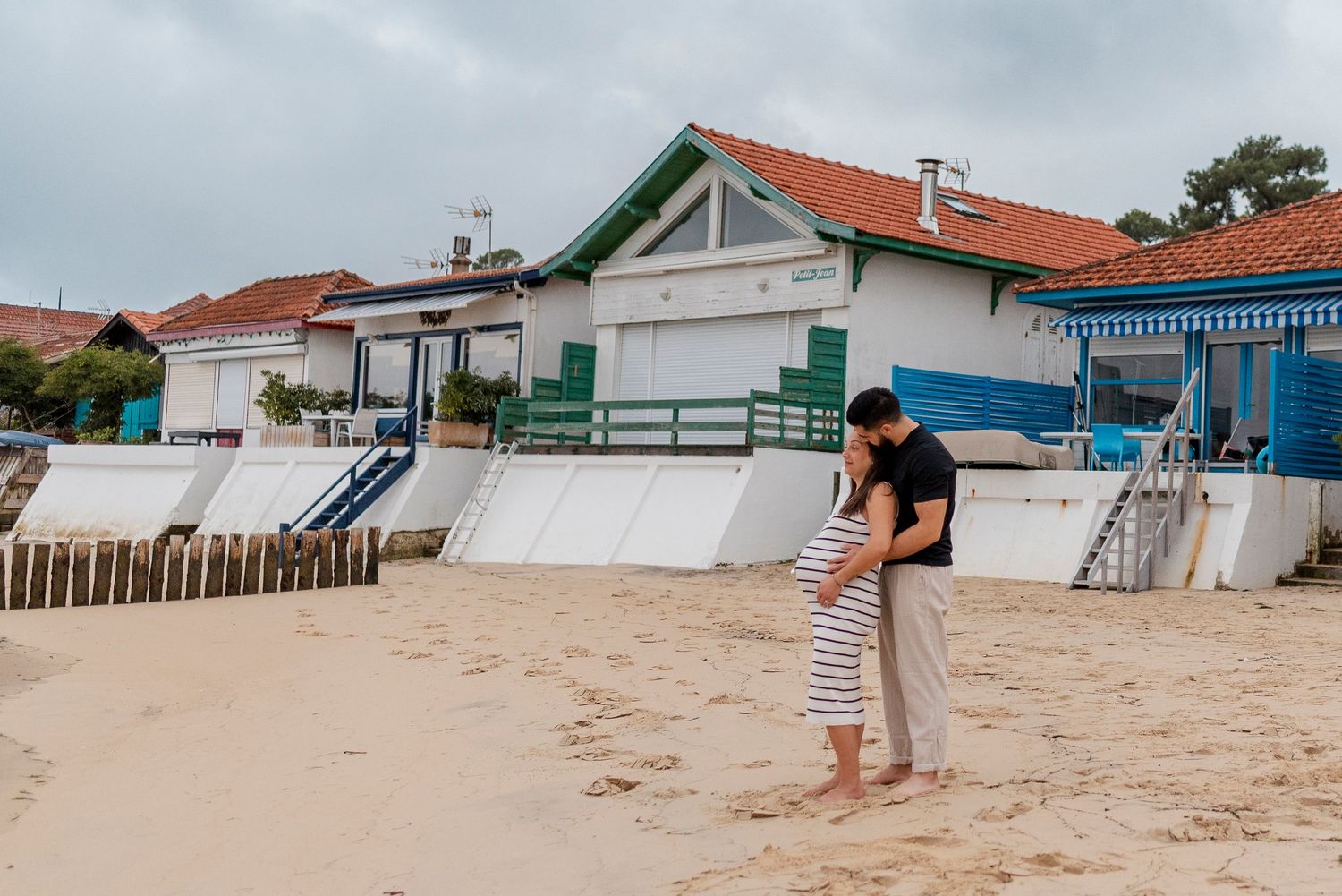 Couple embracing on sandy beach with colorful beach houses in background on cloudy day.