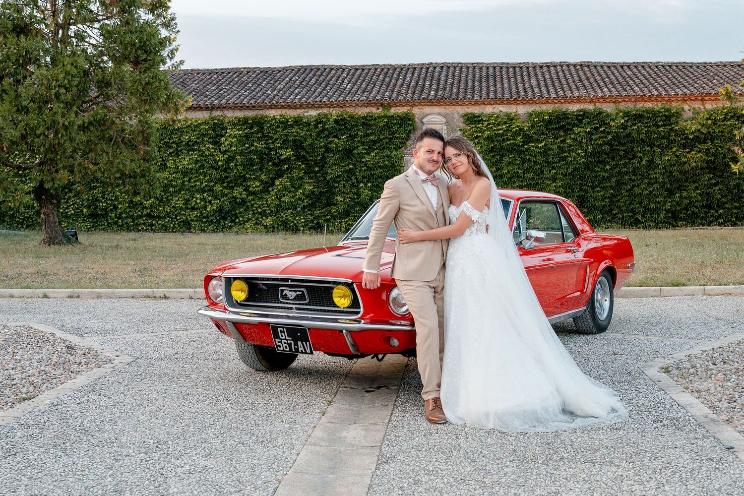 Bride and groom posing together leaning against a classic red Ford Mustang at an outdoor wedding venue.