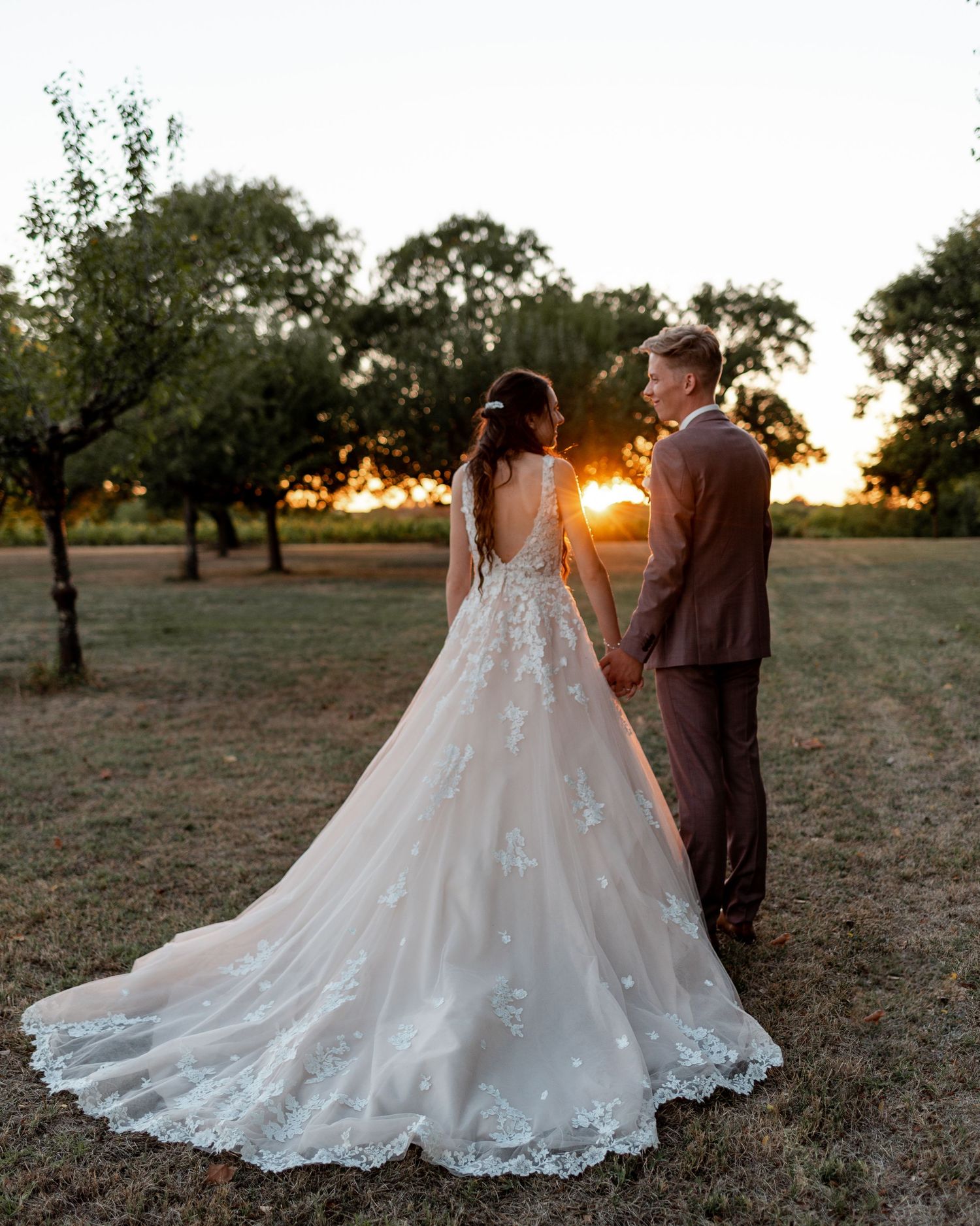 Bride and groom walking through orchard at sunset with dramatic wedding dress train.