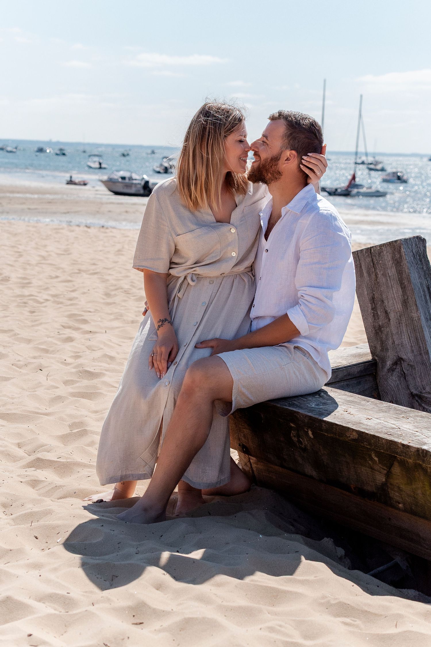 Couple sharing a romantic moment on a wooden beach bench, with sandy shore and sailboats in the background.
