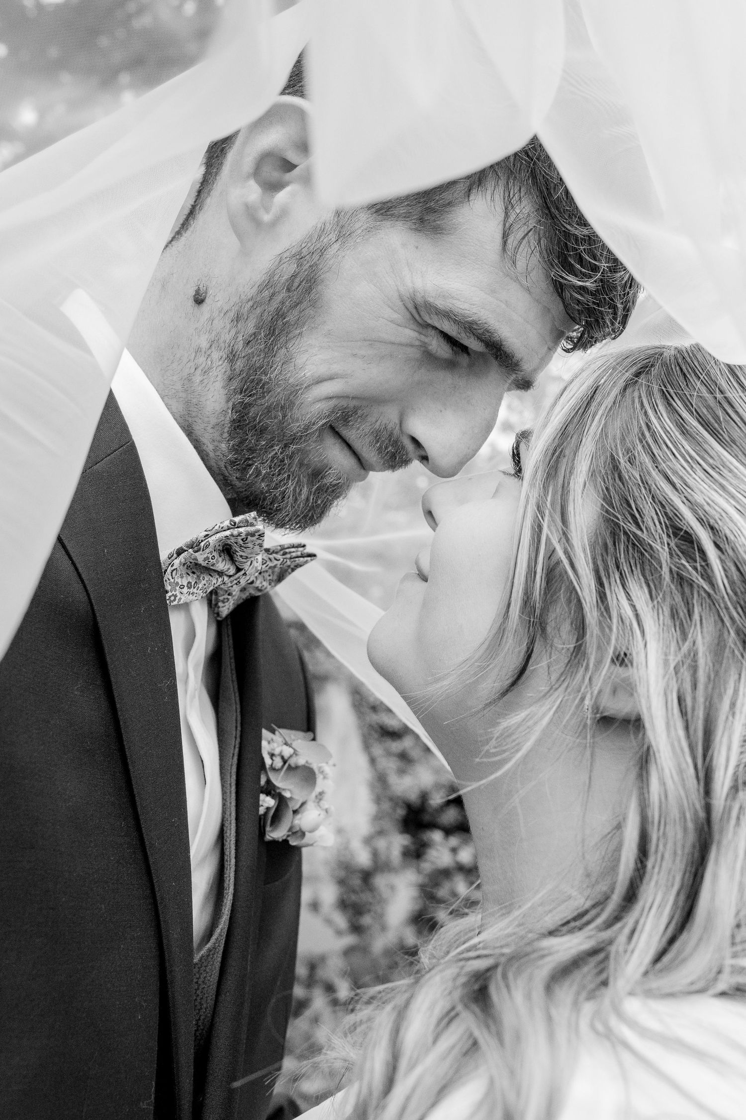 Black and white wedding photo of a smiling groom in a suit and bow tie touching noses with his bride beneath a sheer veil.