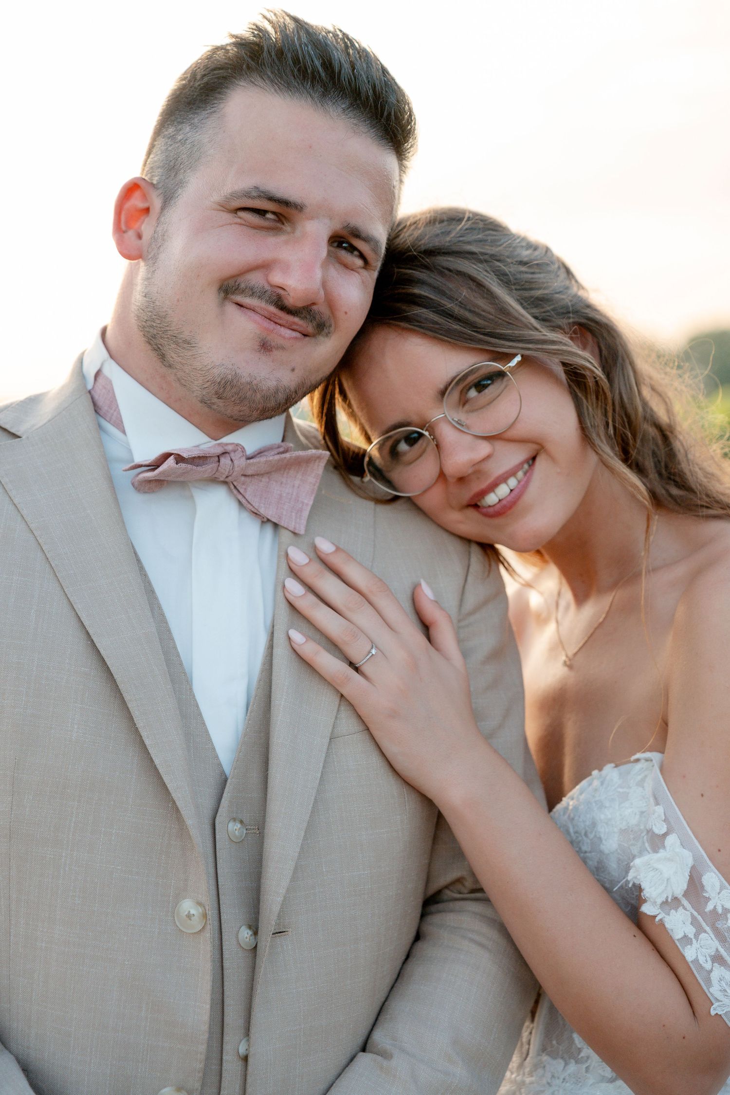 A smiling bride in lace dress and glasses leans on groom in tan suit with pink bow tie during golden hour.